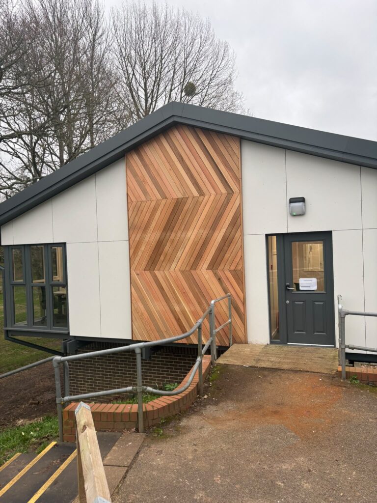 Modern building entrance featuring a distinctive wooden accent wall and accessible ramp surrounded by trees.