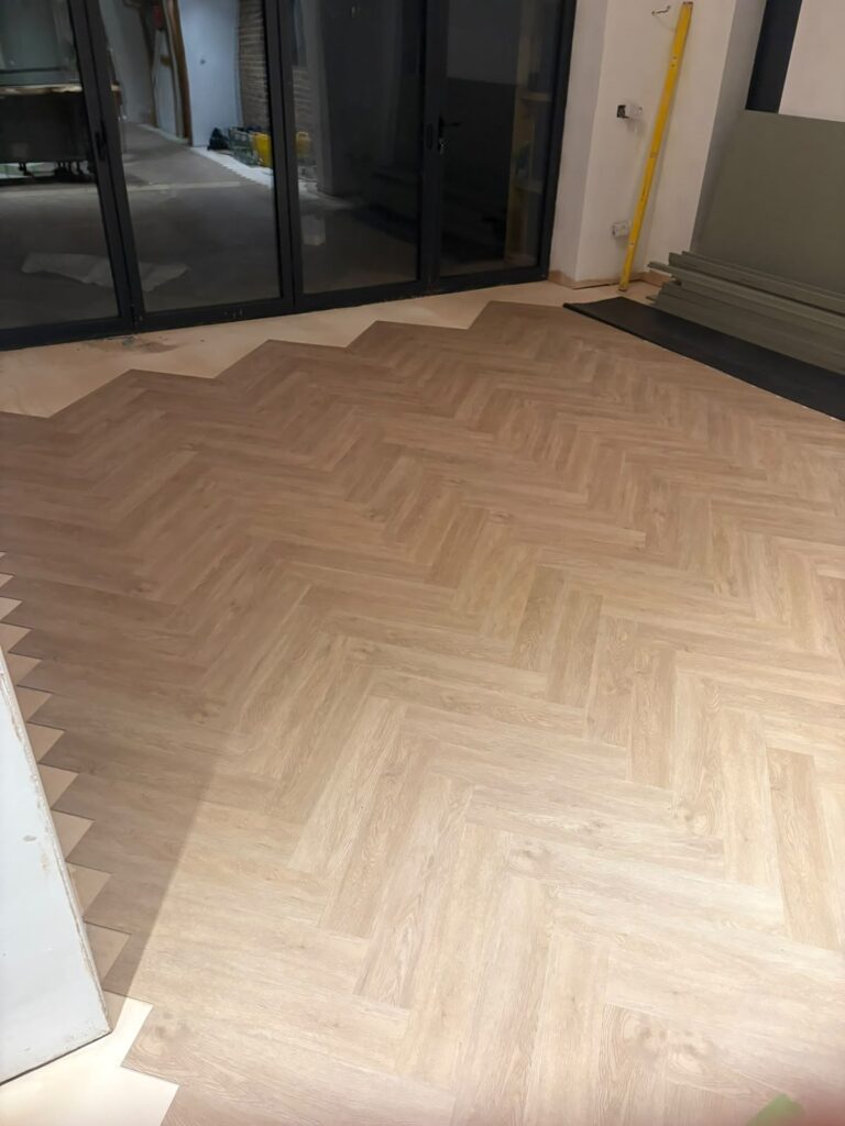 Here are a few concise and accurate alternative text options for the image, depending on the desired level of detail:

*   **Concise:** A newly installed light wood herringbone floor in a room under renovation, with black glass doors and