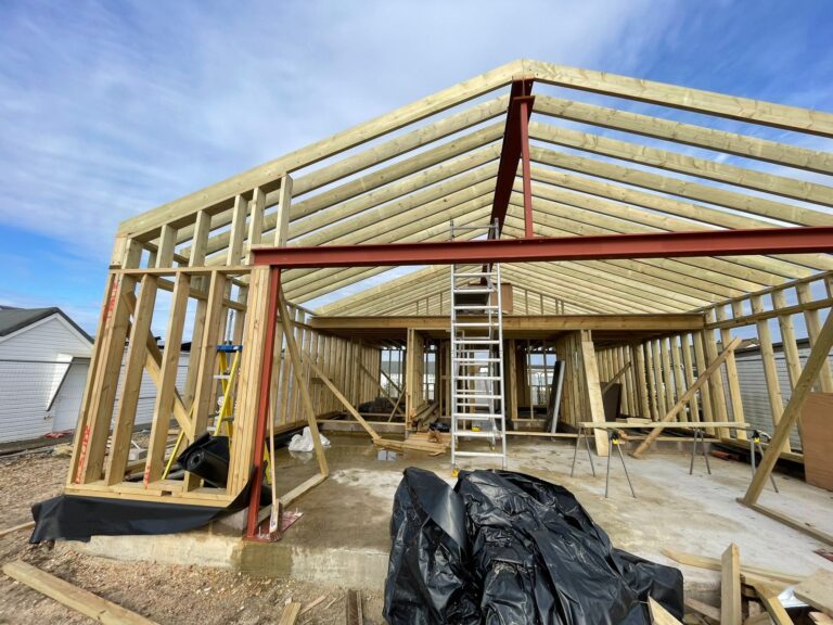 Construction site showing a partially framed house with wooden beams, a metal support structure, and construction materials.