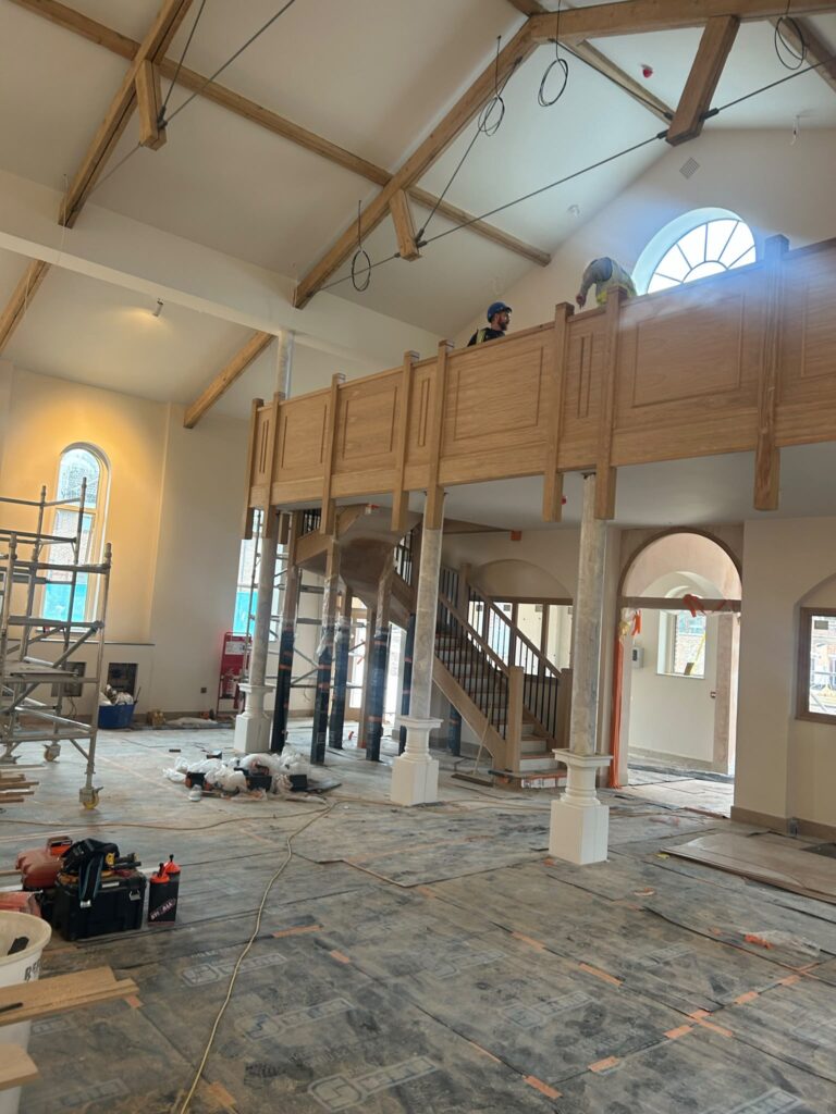 This image displays an interior construction project in a large, open-plan space with high, vaulted ceilings. The room features exposed wooden trusses and beams, some of which are supported by steel cables with hanging loops, possibly for lighting or future fixtures.